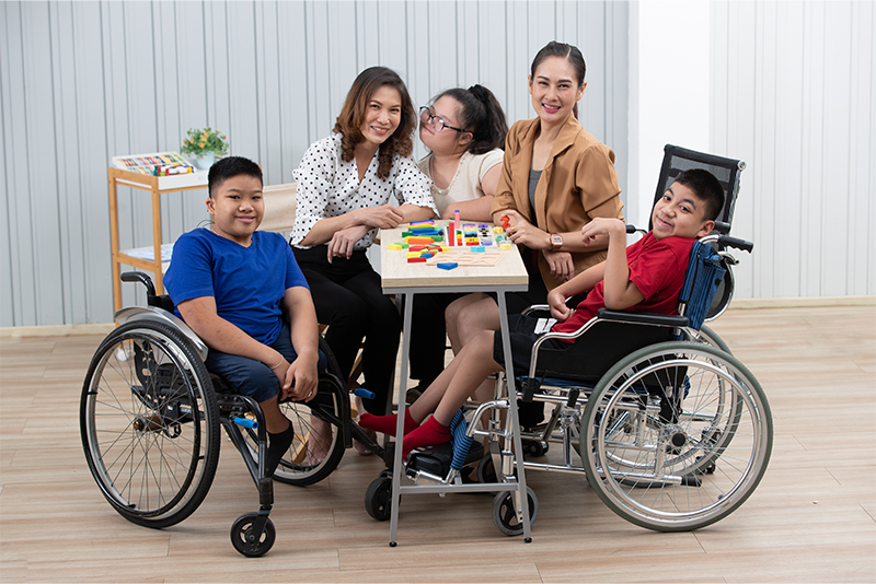 Two adults and three children, two of whom use wheelchairs, are sitting around a table playing with colorful blocks in a bright room.