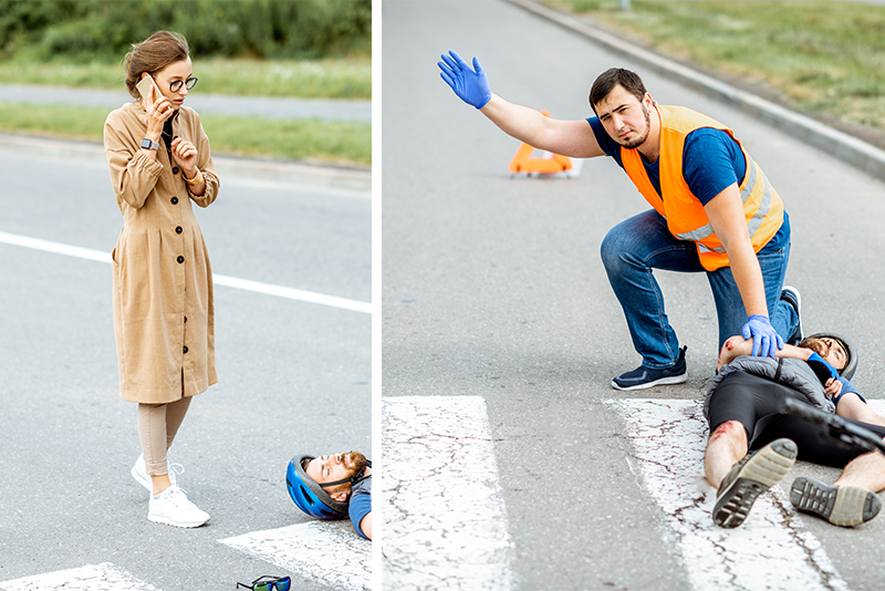 An injured bicyclist in a helmet lies on the street in a crosswalk with a dented car and his broken bike behind him. A man in an orange reflective vest and wearing nitrile gloves is leaning over and touching the victim on the shoulder and waving to get someone to call 911. A woman bystander is calling 911 on her mobile phone.