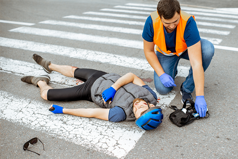 An injured bicyclist in a helmet lies on the street in a crosswalk with a dented car and his broken bike behind him. A man in an orange reflective vest and wearing nitrile gloves is leaning over and touching the victim on the shoulder. A first aid kit is open next to the man as he is rendering aid to the victim.