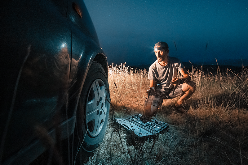 A man is changing the tire of his car on the side of the roadin the dark. He is wearing a headlamp.