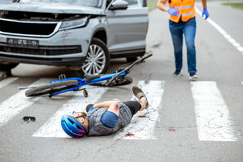 An injured bicyclist in a helmet lies on the street in a crosswalk with a dented car and his broken bike behind him. A man in an orange reflective vest is running to the scene of the accident.