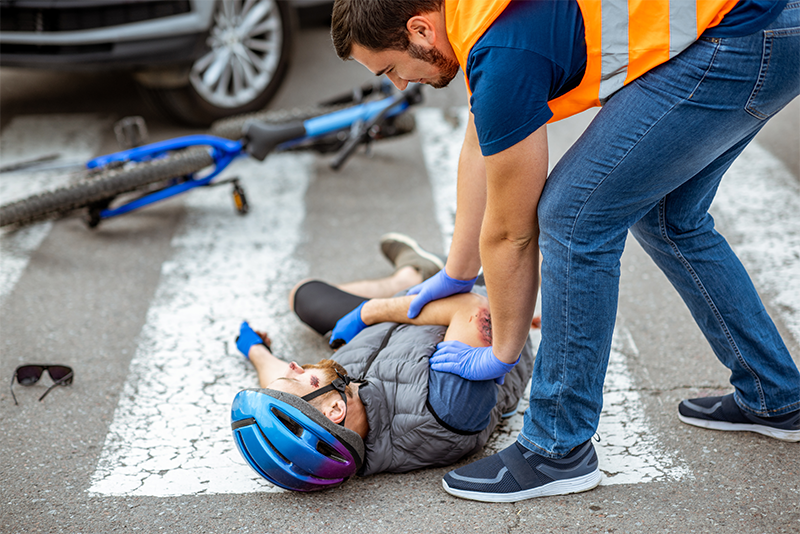 An injured bicyclist in a helmet lies on the street in a crosswalk with a dented car and his broken bike behind him. A man in an orange reflective vest and wearing nitrile gloves is leaning over and touching the victim on the shoulder.