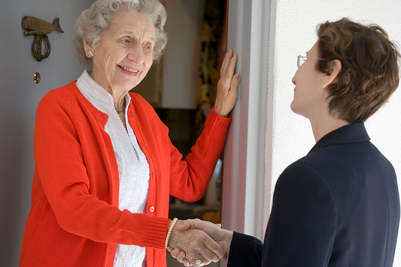A senior woman wearing a red cardigan and white blouse is smiling and shaking hands with a younger woman in a dark blazer at a doorway.