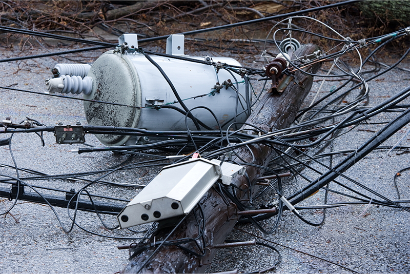 High power electric lines and a wooden power pole that have fallen onto an asphalt road.