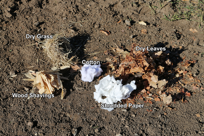 Small piles of dry grass, wood shavings, cotton, shredded paper, and dry leaves on dry dirt.