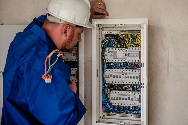 An electrician in a white hardhat and a blue jacket looks at the electrical paneling on a wall.