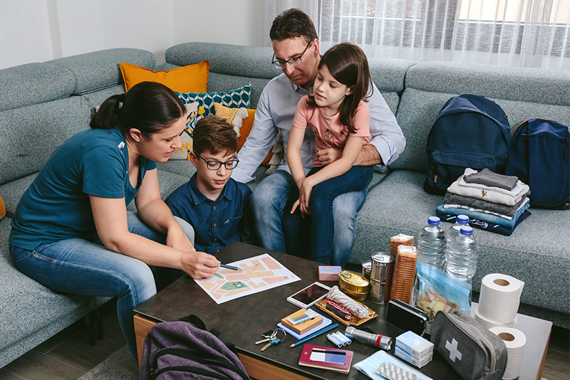 A family of four sitting on a couch and floor are engaged in planning or discussing an emergency preparedness plan map on a coffee table surrounded by various supplies including water bottles, snacks, and first aid items.