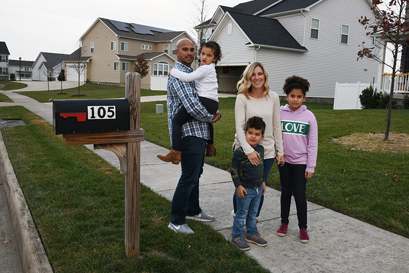 A family of five people, including three children and two adults, stand on a sidewalk in a suburban neighborhood near a mailbox labeled 105.