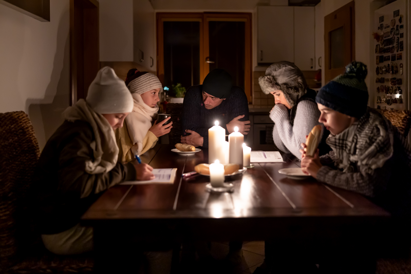 A family bundled in warm clothing sits in the dark around a dining table with a candle in the middle.