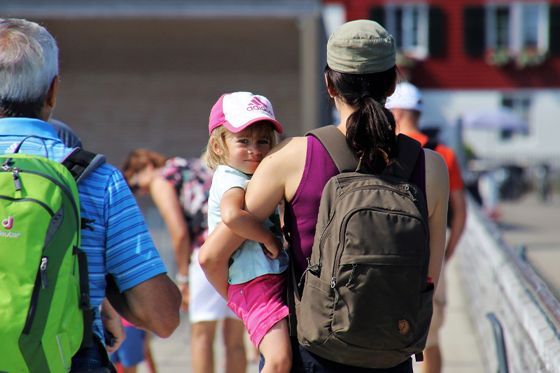 A woman wearing a hat and backpack is carrying a child dressed in a pink outfit and white cap while walking outdoors among other people.