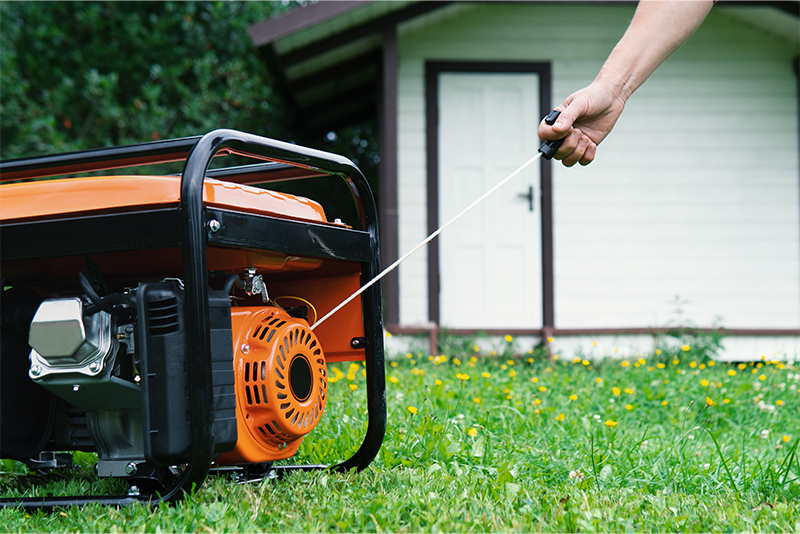 An orange and black gas powered generator sits on a backyard lawn. A hand is pulling on the starter rope.
