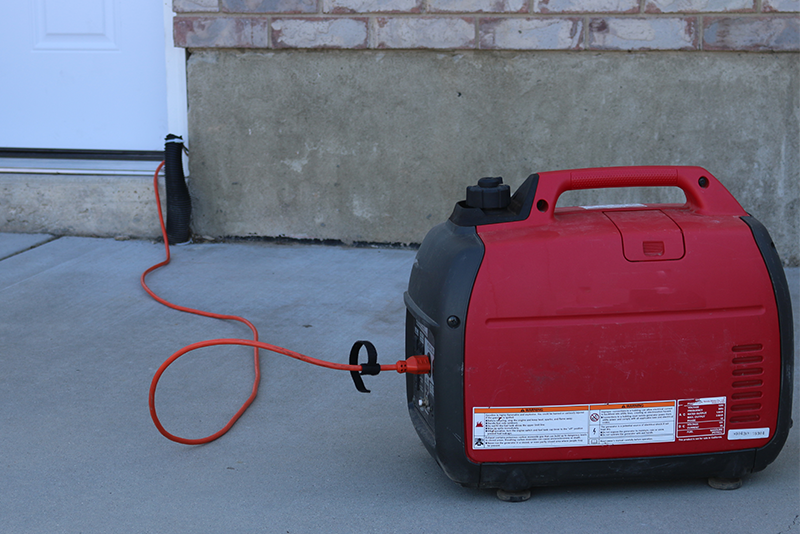 A red 2000 watt gas generator runs outside on a patio. An electrical cord is plugged into the generator and runs up to the house and in through a door.