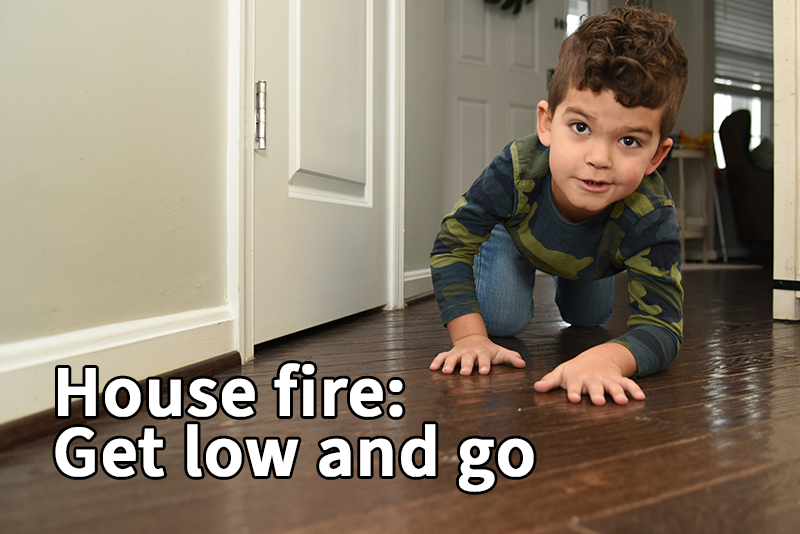 A child wearing a camouflage long-sleeve shirt and jeans is crawling low on a wooden floor near a door with the text advising to get low and go during a house fire.