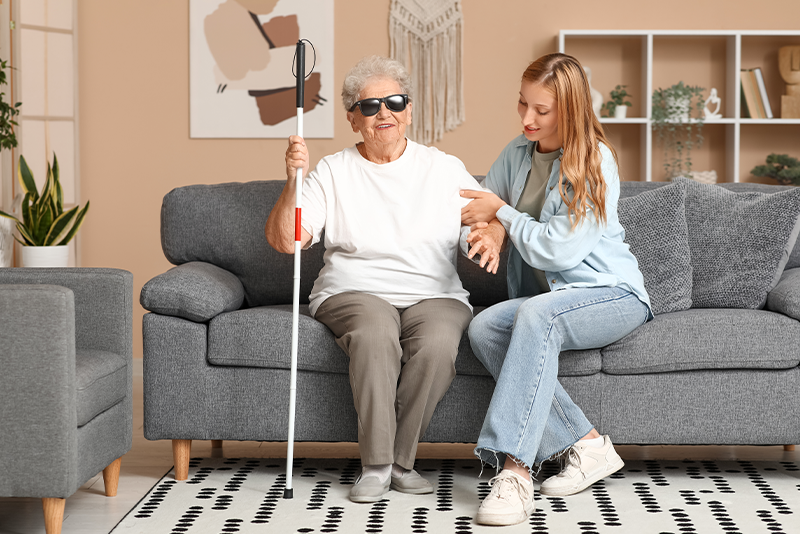 A senior blind woman wearing sunglasses and holding a white cane is sitting on a gray couch while a younger woman in casual clothes sits beside her, offering support.
