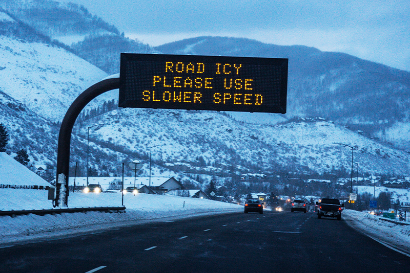 A snowy mountain road with several cars driving under a digital sign warning that the road is icy and advising slower speeds.