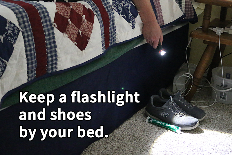 A man's hand holds a powered on flashlight projecting a beam of light onto a carpeted floor next to a bed. A pair of shoes and a chemical light stick are also on the floor. Text reads: "Keep a flashlight and shoes by your bed."