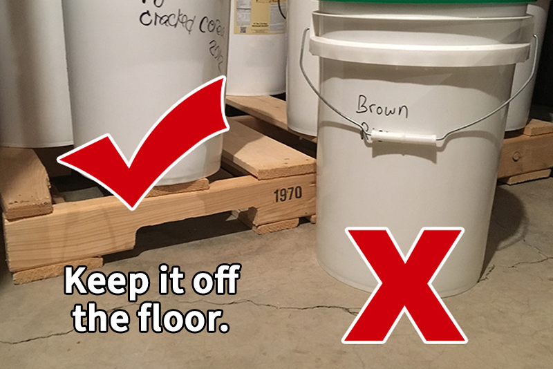 A food storage plastic bucket sitting on a concrete floor with an "X" over it. Another bucket sits on a wooden pallet with a check mark on it. Text reads, "Keep it off the floor."