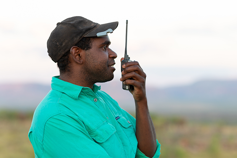 A person wearing a green shirt and a black cap is holding and speaking into a walkie-talkie outdoors with a blurred natural landscape in the background.