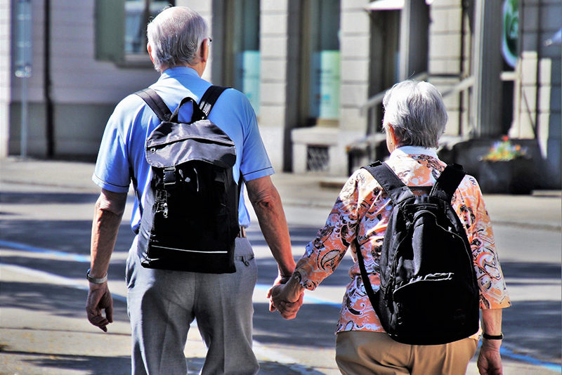 Two seniors wearing backpacks are walking hand in hand on a city street in bright daylight.
