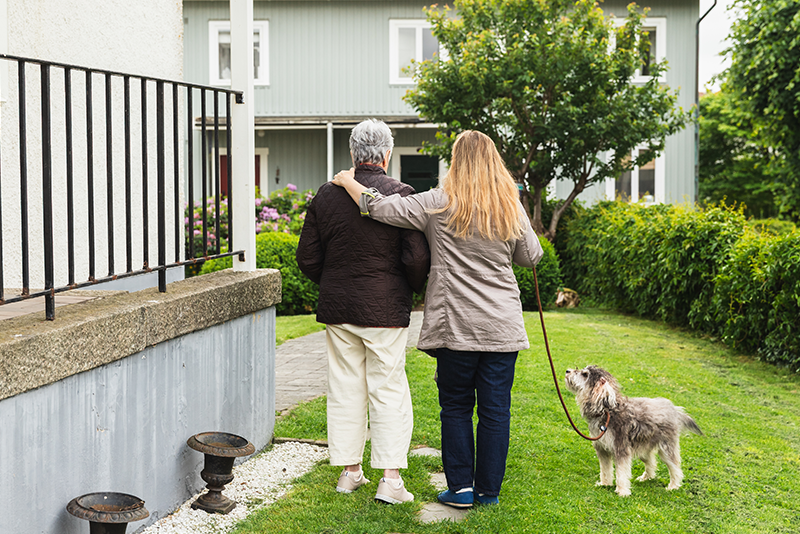 Two adults walk together on a grassy path near a house, with one adult holding a small dog on a leash.
