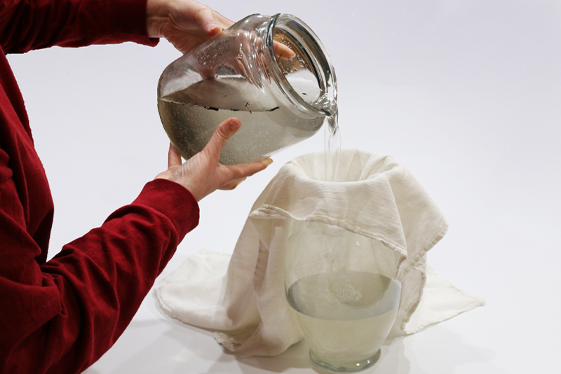 Woman pouring dirty water from a large glass jar through a cloth and into another glass jar.