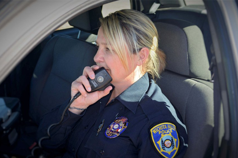 A woman in a police uniform is sitting in a vehicle and speaking into a handheld radio.