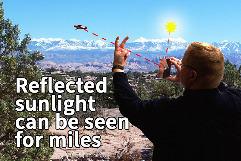 A person outdoors uses a handheld mirror to reflect sunlight through his upheld fingers, toward a small red airplane, with text explaining that reflected sunlight can be seen for miles.