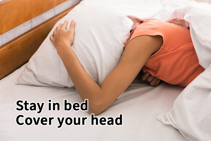 A woman lying face down in bed with an arm and pillow over her head is practicing the protective action for earthquake if your are in bed. Text reads, "Stay in bed. Cover your head."