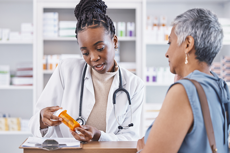 A woman in a white coat with a stethoscope is explaining medication from a prescription bottle to an older woman across a counter in a pharmacy setting.