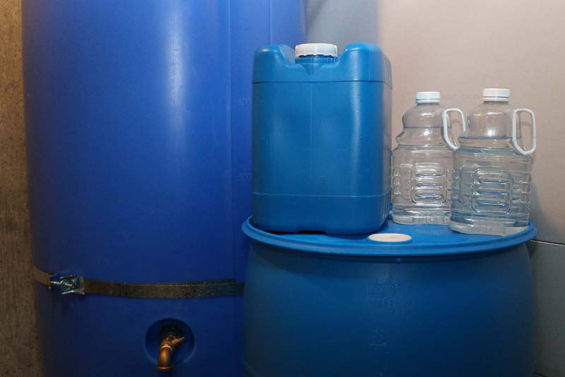 A 250 gallon water storage container next to a 55 gallon water storage container, a 5 gallon jug, and two  juice bottles used for storing water.