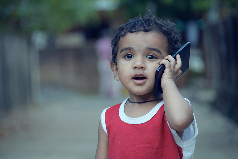 A young boy in a red shirt is speaking on a cell phone while outdoors.